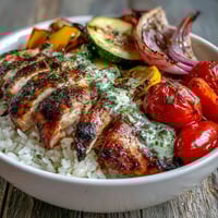 Golden-brown Lemon Herb Roasted Chicken Breast slices rest on fluffy white rice, next to vibrant red bell peppers and zucchini. The bowl is finished with a drizzle of zesty lemon dressing.